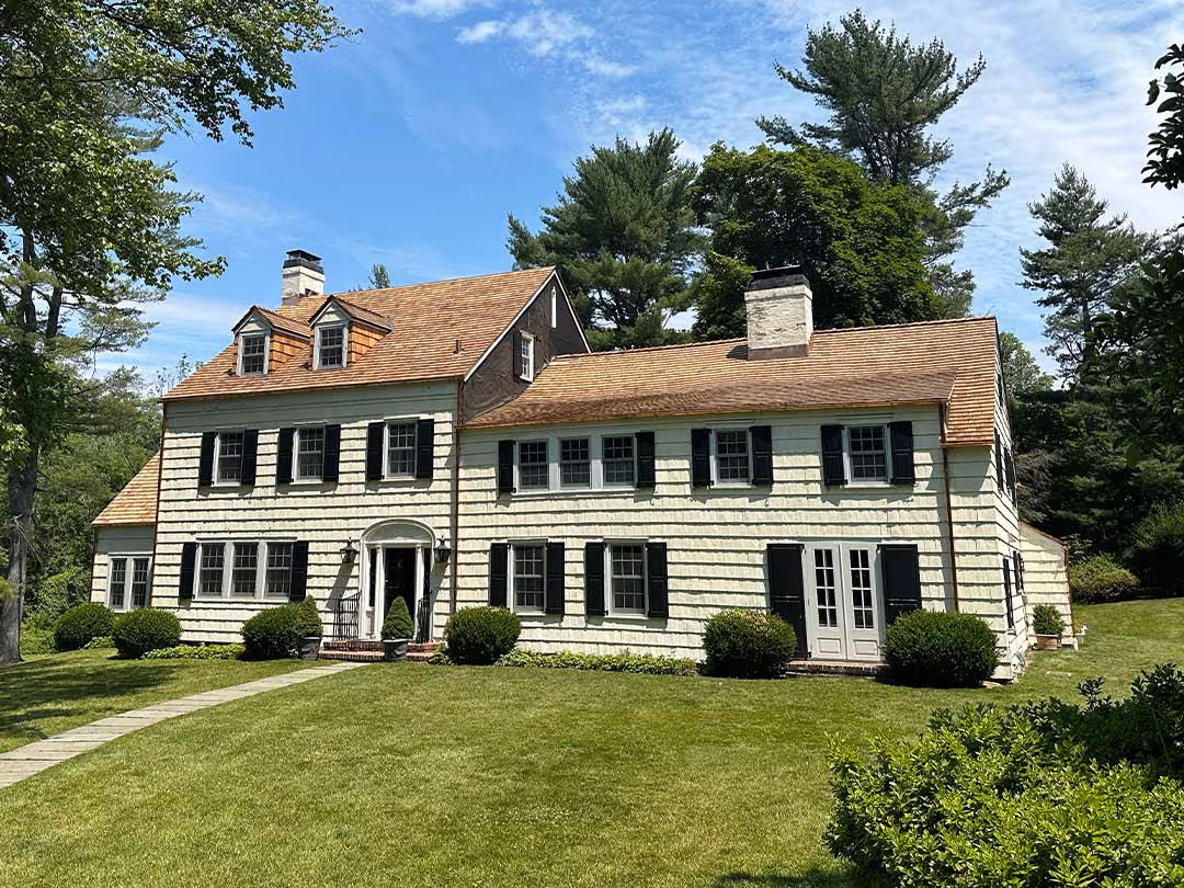 Two-story house with cedar roof shingles, white exterior, black shutters, and green lawn under blue sky.