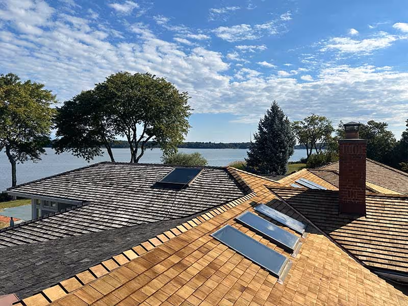 New wooden shingle roof with skylights and a chimney overlooking a lake and trees.