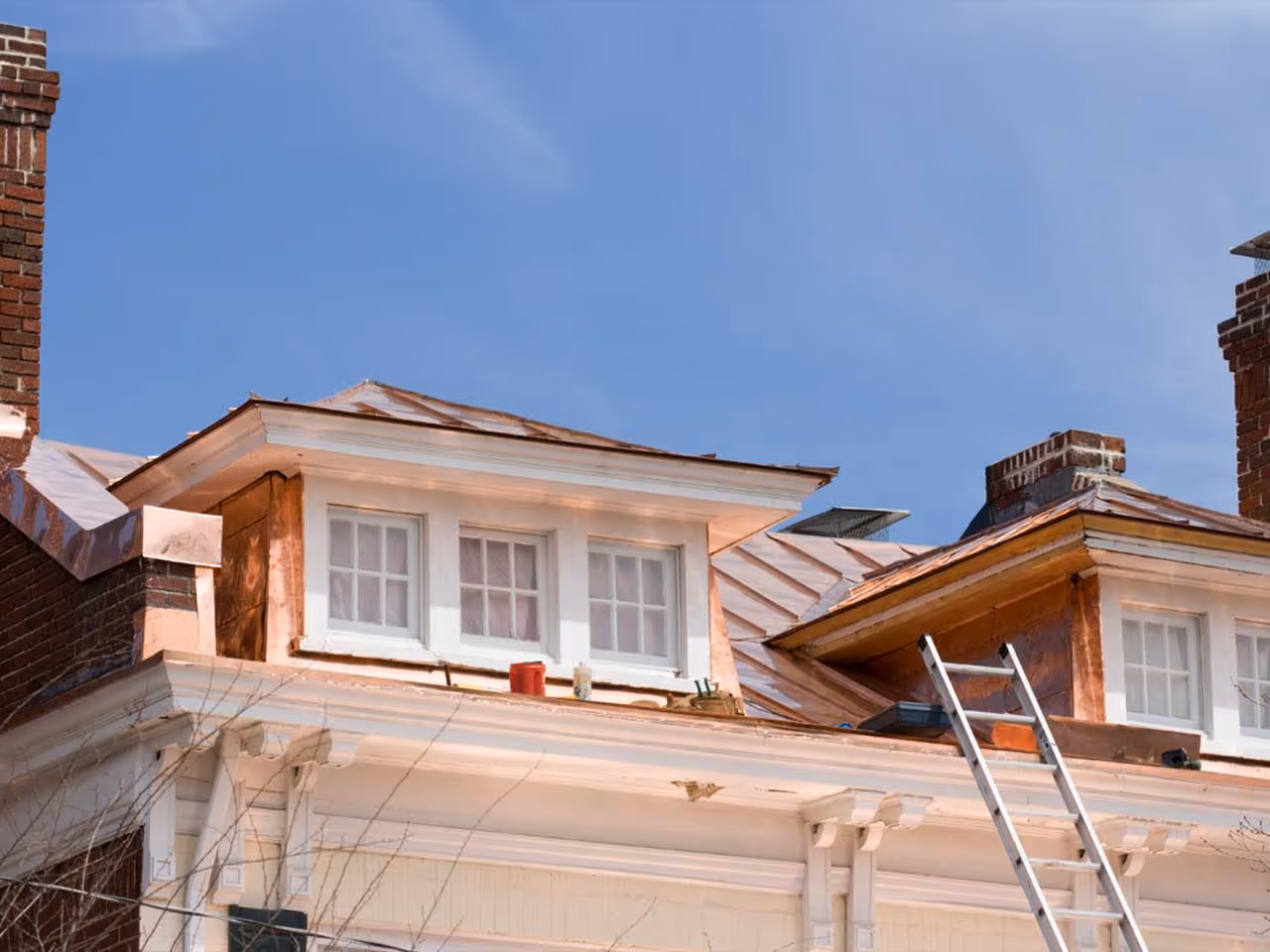 Copper metal roofing on dormer windows of a white house under clear blue sky with a ladder leaning on the roof.