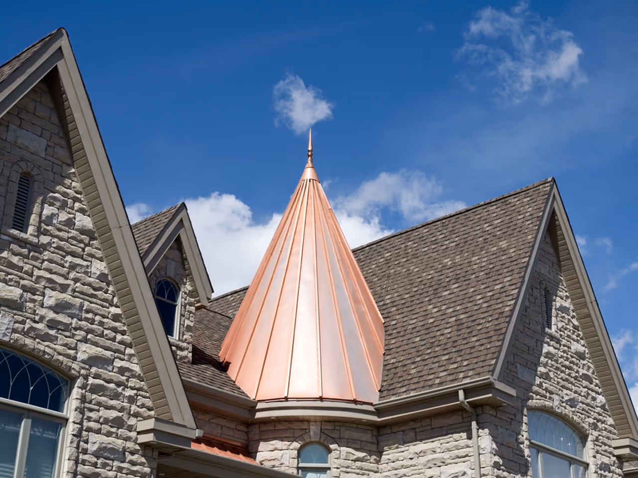 Stone house with a steep gray shingle roof and a pointed copper metal turret under blue sky.