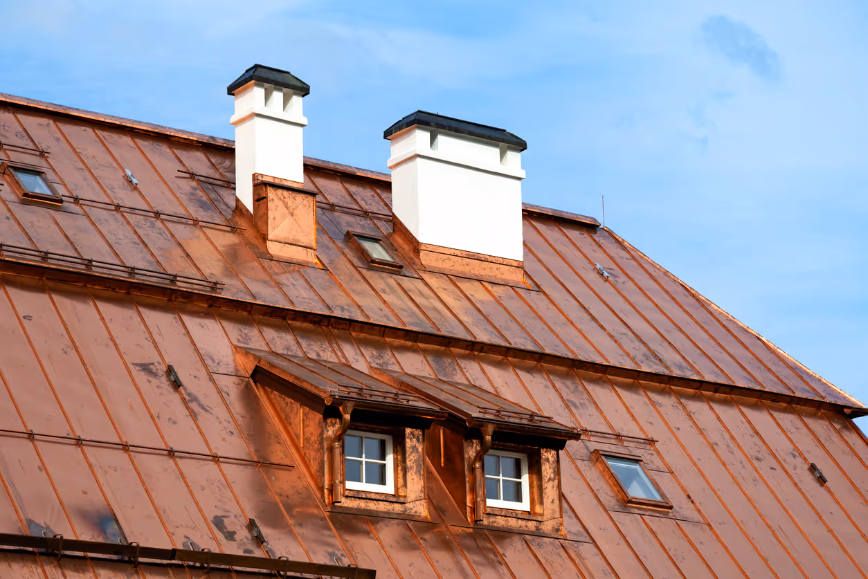 Copper metal roof with two white chimneys and two small dormer windows under blue sky.