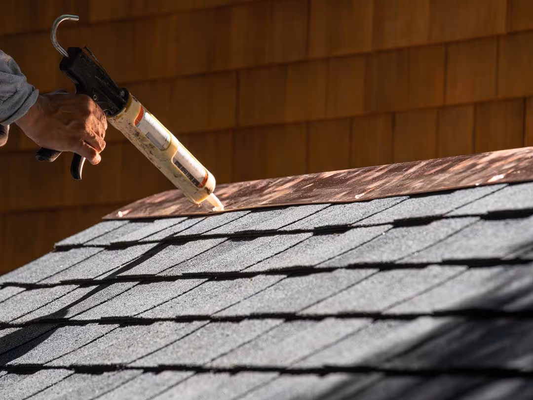 Hand holding a caulking gun applying adhesive along the edge of copper flashing on an asphalt shingle roof.