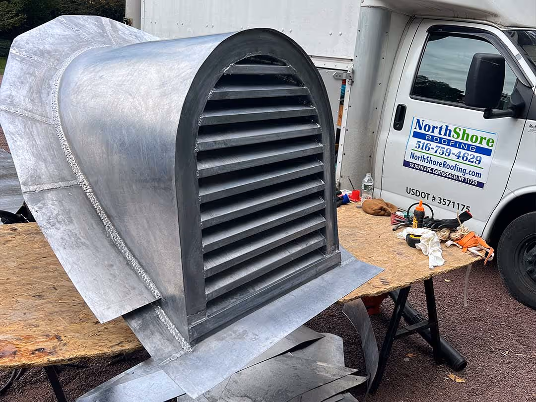Large galvanized metal roof vent resting on a wooden table with a roofing company van in the background.
