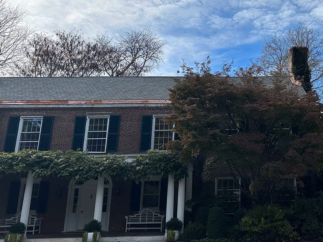 Two-story brick house with white columns, black shutters, benches, and surrounding greenery under a partly cloudy sky.