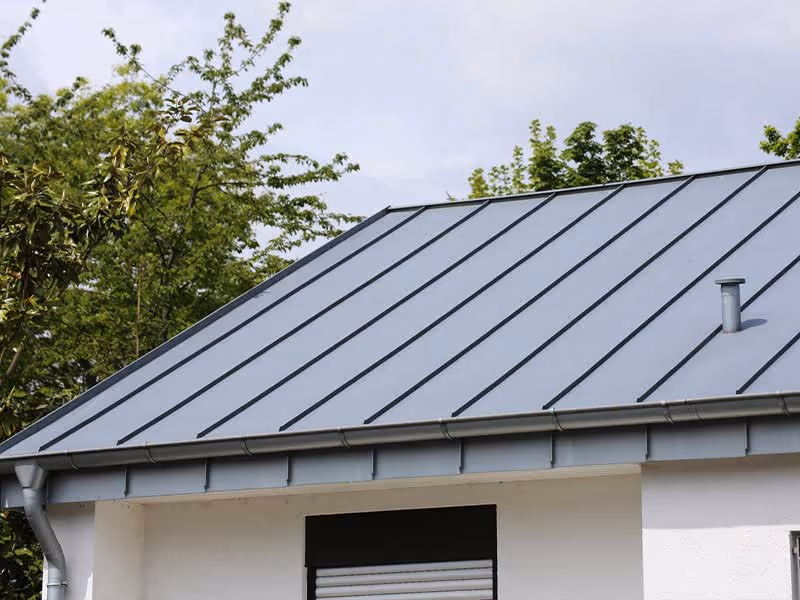 Close-up of a modern gray metal roof with vertical seams on a white house under a cloudy sky.
