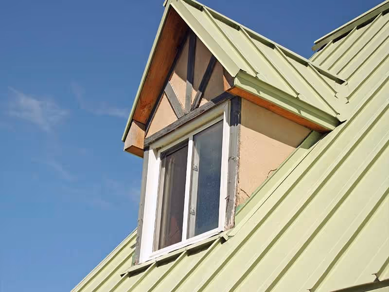 Close-up of a green metal roof with a dormer window under a clear blue sky.