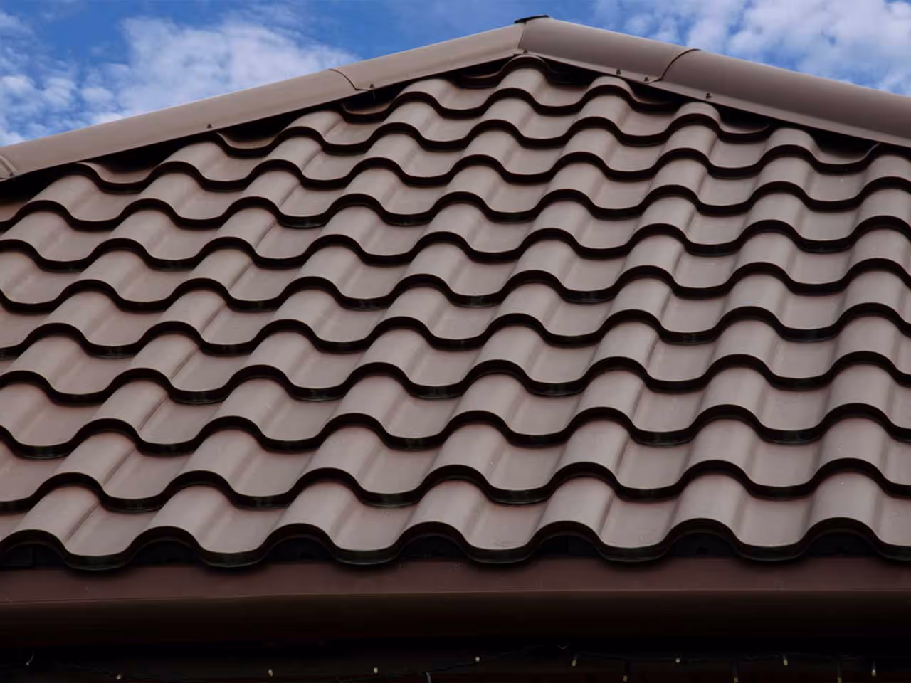 Close-up of brown metal roofing tiles with a wavy pattern under a blue sky.