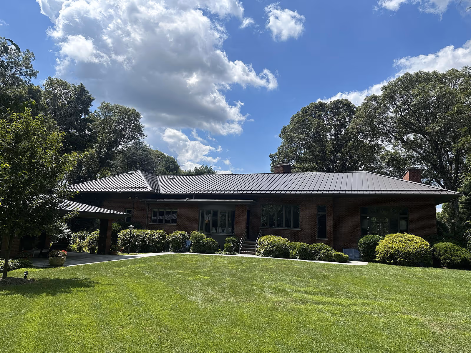 Single-story brick house with a dark metal roof surrounded by green trees and lawn under a partly cloudy sky.