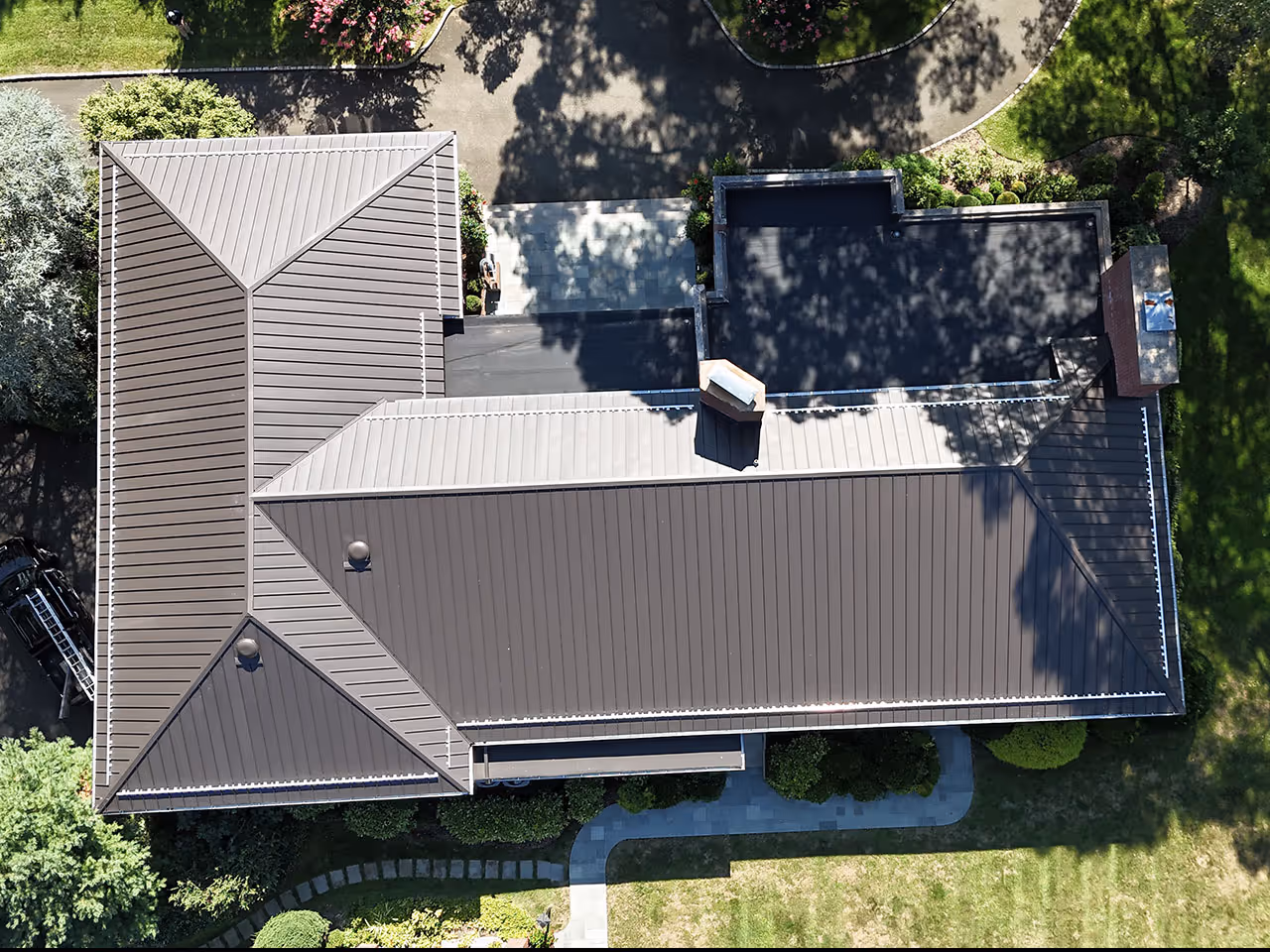 Aerial view of a house with a large brown metal roof surrounded by green trees and a manicured lawn.