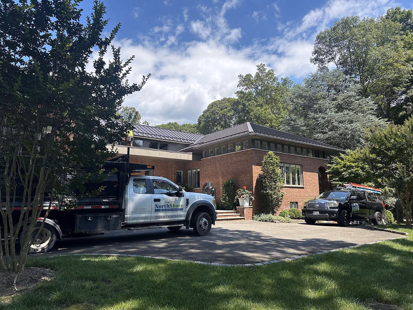 Brick house with metal roofing and two parked work trucks in a driveway, surrounded by trees.