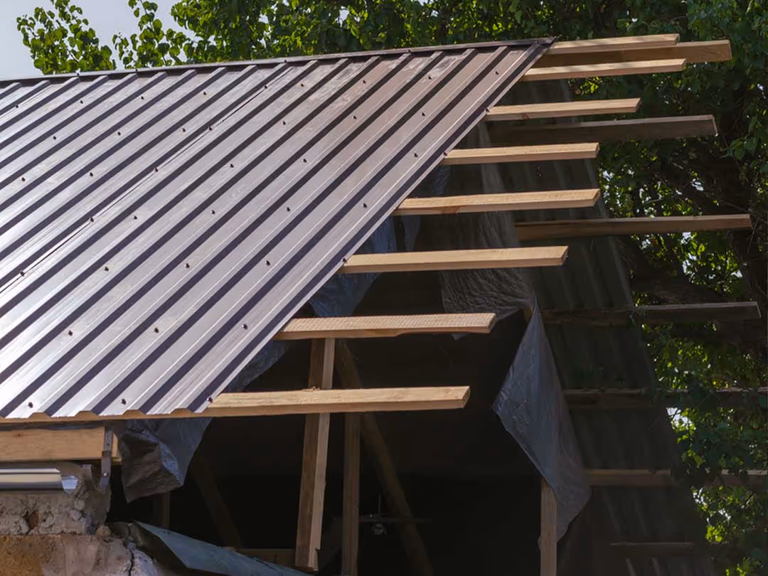 Partially installed brown metal roofing on wooden frame structure with green foliage in background.