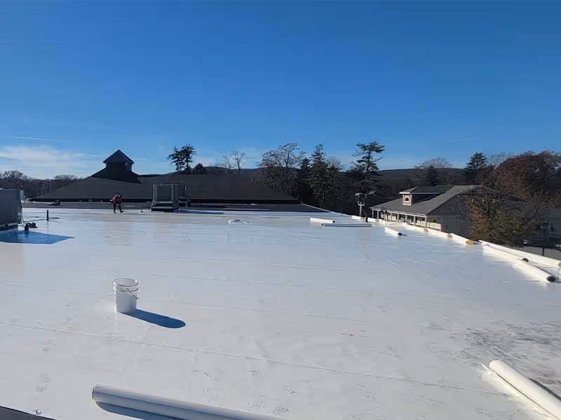 Flat white roofing on a building under clear blue sky with workers and nearby buildings in background.