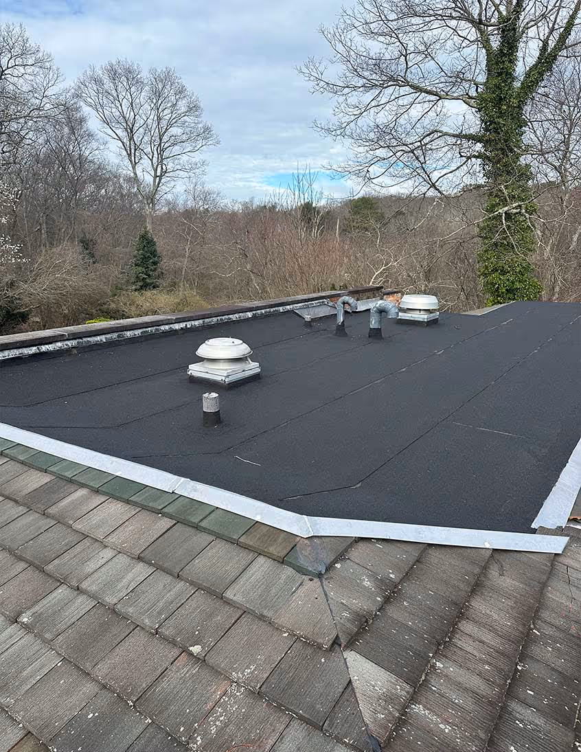 Black flat roofing with vents installed on top of a shingled roof surrounded by bare trees.