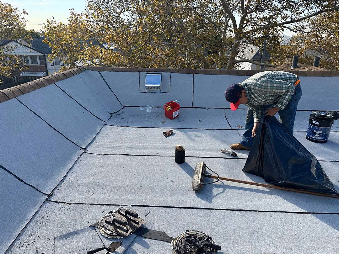 Man wearing a cap and plaid shirt working on flat roof repair with tools and roofing materials around.