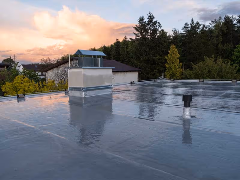 Flat rooftop with reflective waterproof coating at sunset, surrounded by trees and neighboring buildings.