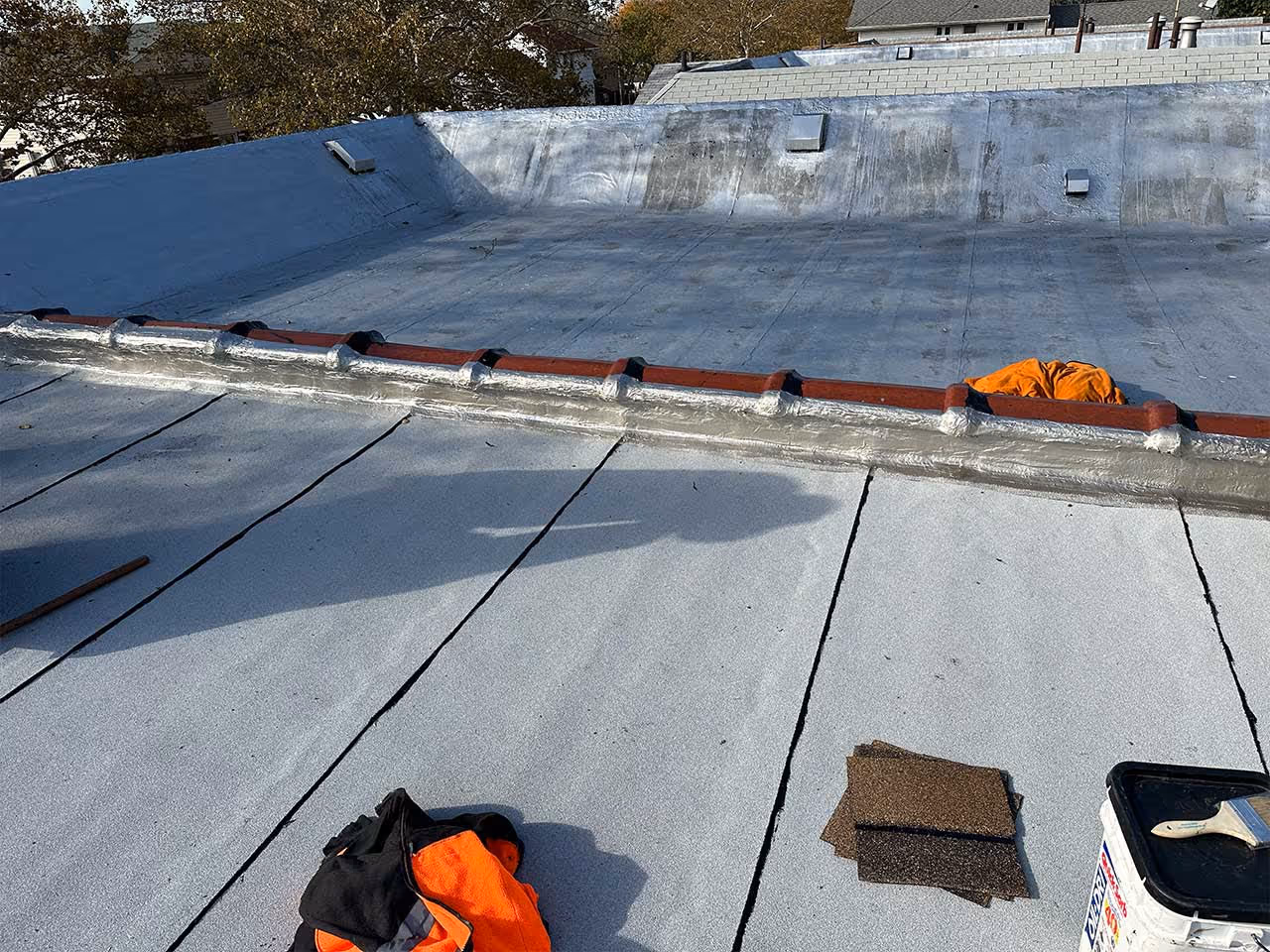 Flat roof with gray waterproofing material, orange clothes, roofing shingle pieces, and a paintbrush on a bucket.