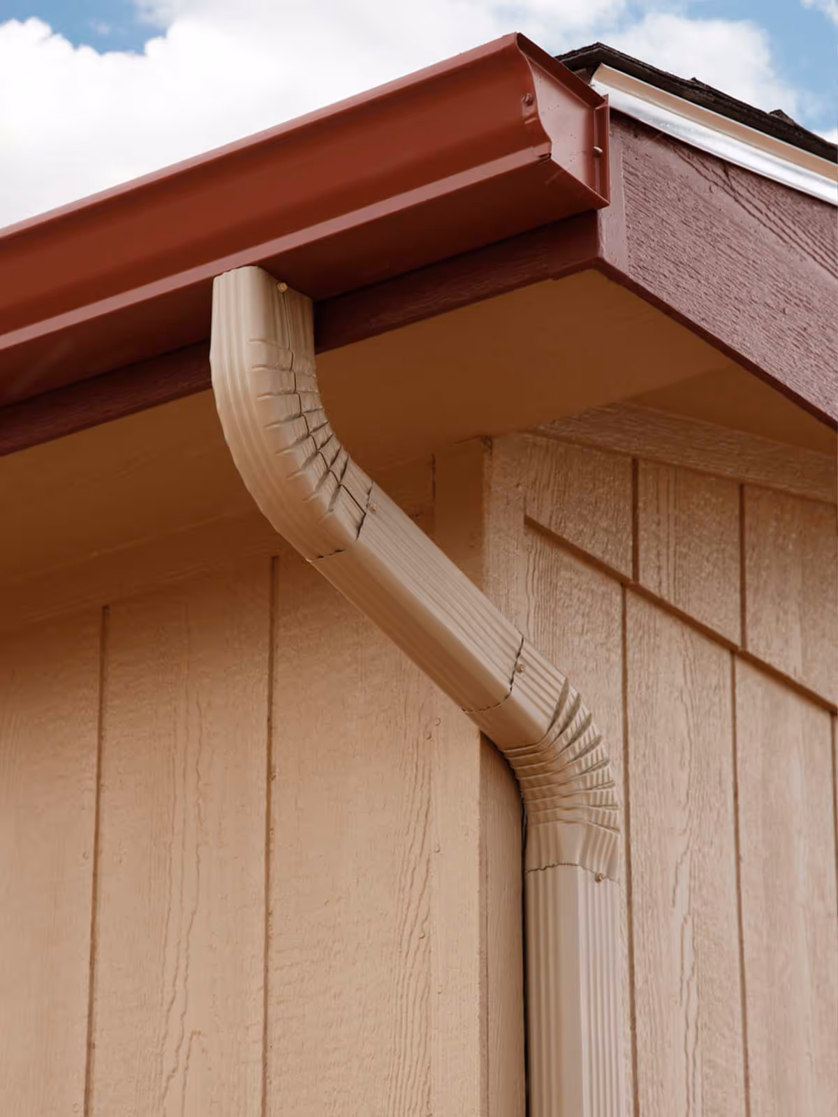 Close-up view of a beige downspout connected to a brown gutter on a wooden house exterior.