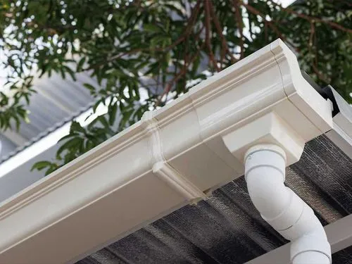 Close-up of white aluminum gutters and downspout on a roof with trees in the background.