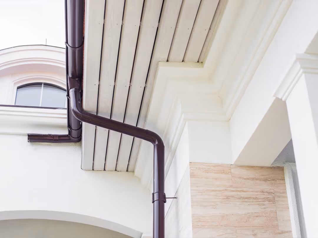 Black rain gutter and downspout attached to the roof edge of a white building with beige stone wall.