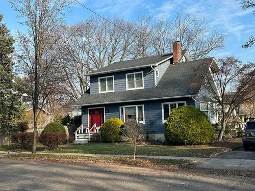 Two-story blue house with a red front door, surrounded by leafless trees and bushes under a partly cloudy sky.