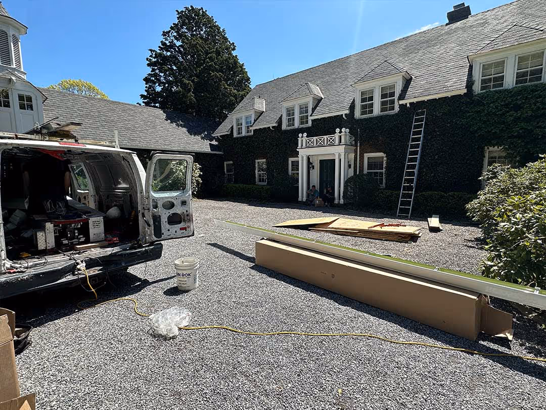 Work van with open rear doors and tools next to large brick house with ivy and a ladder on gravel driveway.