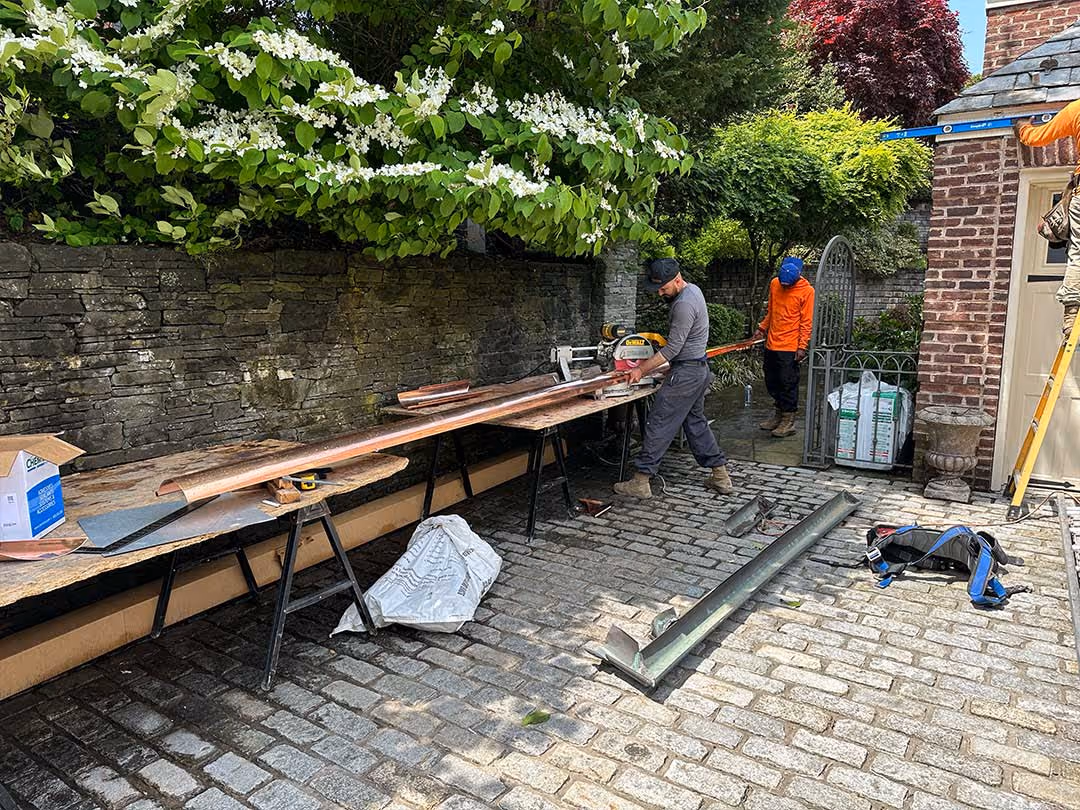 Two workers handling long metal gutters in a paved courtyard next to a brick building and stone wall.