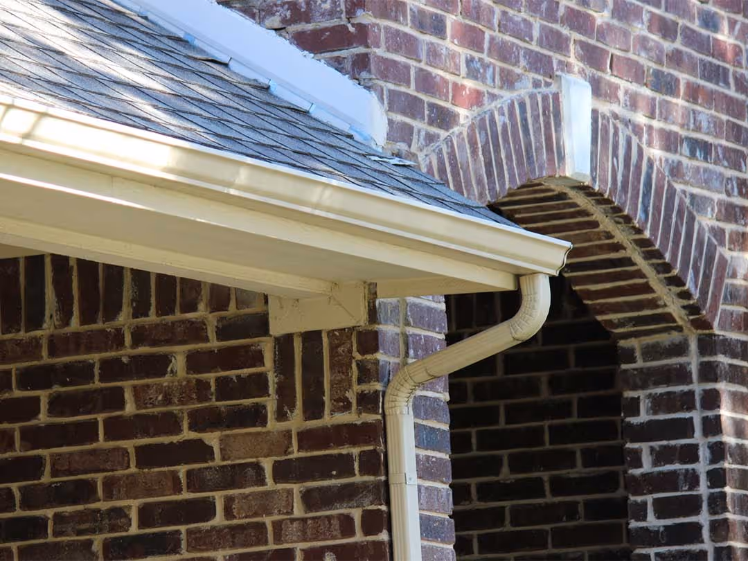 White aluminum gutter and downspout on brick house with shingled roof.