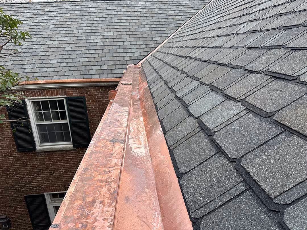 Copper rain gutter installed alongside shingled roof on a brick house.