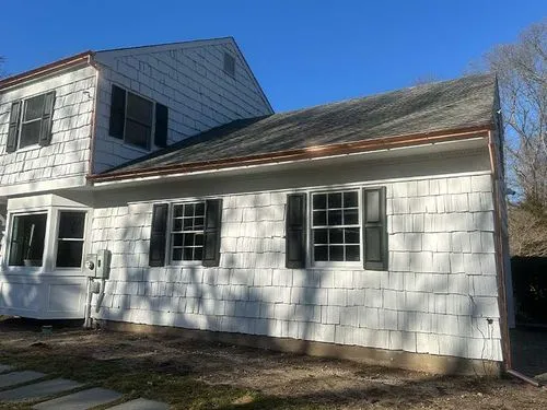 White two-story house with black shutters and a newly installed copper gutter under a clear blue sky.
