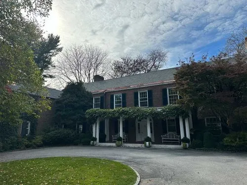 Two-story brick house with white columns, green shutters, and a covered porch with a swing under a partly cloudy sky.