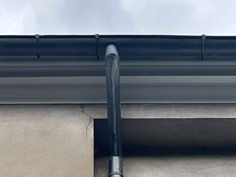 Close-up of a black gutter downspout attached to a beige building wall under a dark gutter.