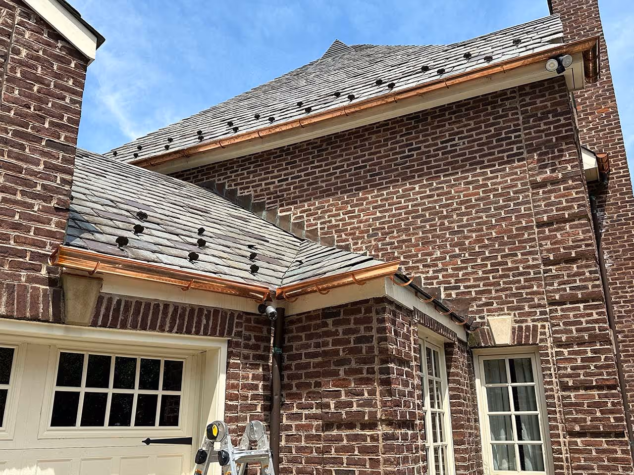 Copper gutters installed along the edges of a brick house roof with slate shingles under blue sky.