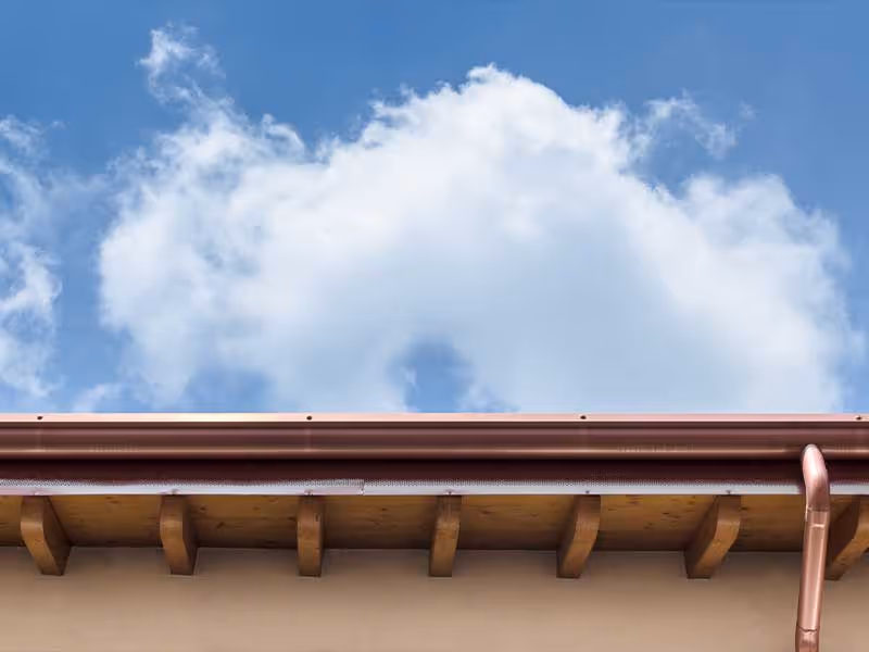 Copper gutter attached to a wooden roof edge under blue sky with white clouds.