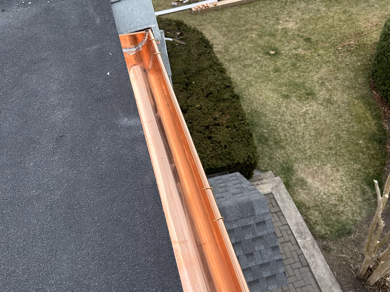 Copper gutter attached to roof edge draining water outside above a green lawn and paved path.