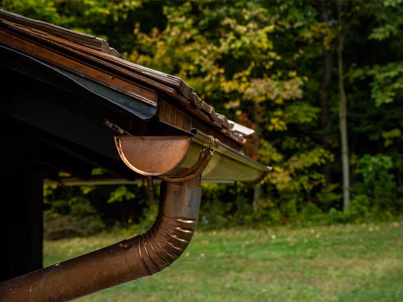 Copper gutter system attached to the edge of a roof with green trees and grass in the background.