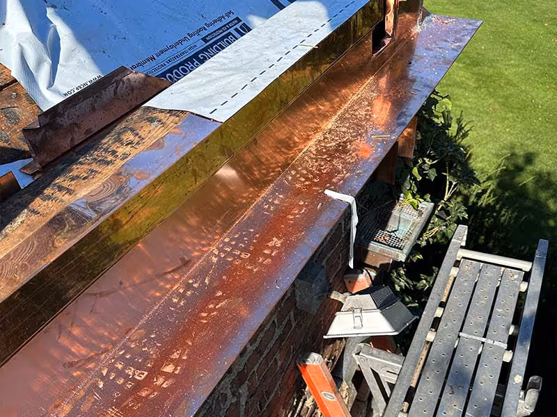 Newly installed shiny copper gutter on a roof with construction materials and a ladder nearby.