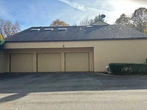 Beige building with dark shingle roof, three closed garage doors, and skylights under a blue sky.