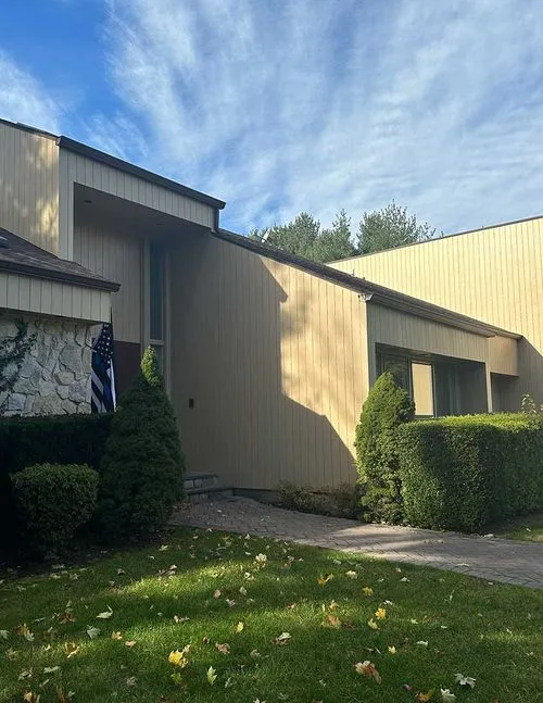 Modern house entrance with beige vertical siding, trimmed bushes, and a stone wall under a partly cloudy sky.