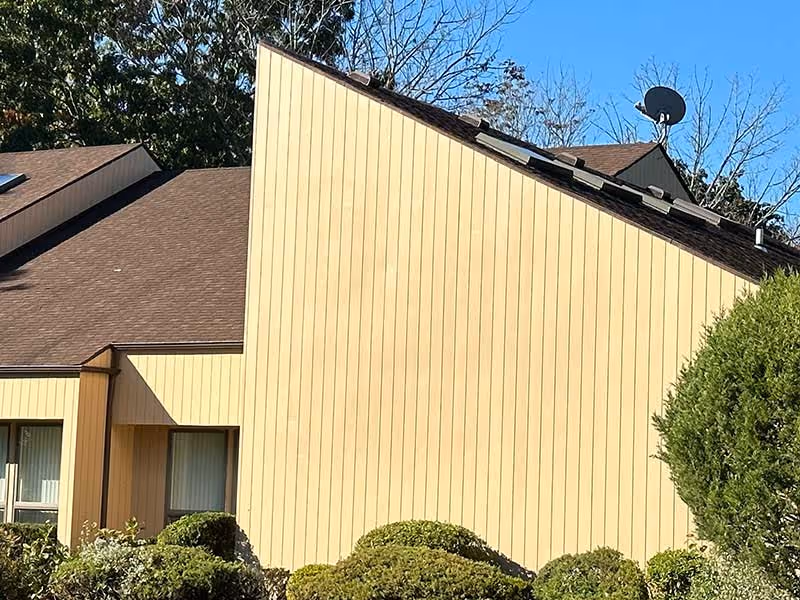 Yellow vertical cedar siding on house with brown roof, surrounded by bushes under clear blue sky.