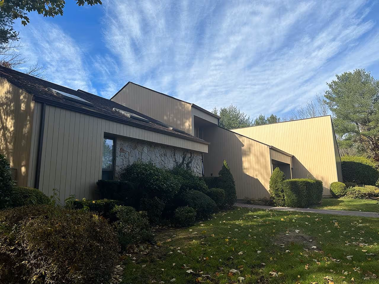 Modern house with beige siding, stone accent, skylights, and well-trimmed shrubs under a blue sky.