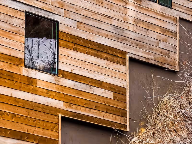 Partially completed cedar wood siding on building exterior with window and exposed sheathing below.