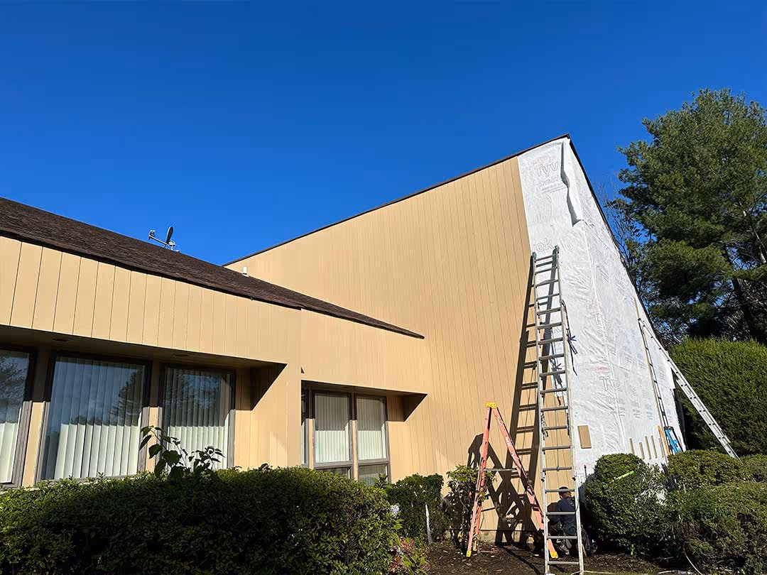 Building exterior with beige vertical siding partially installed and three ladders leaning against the wall.