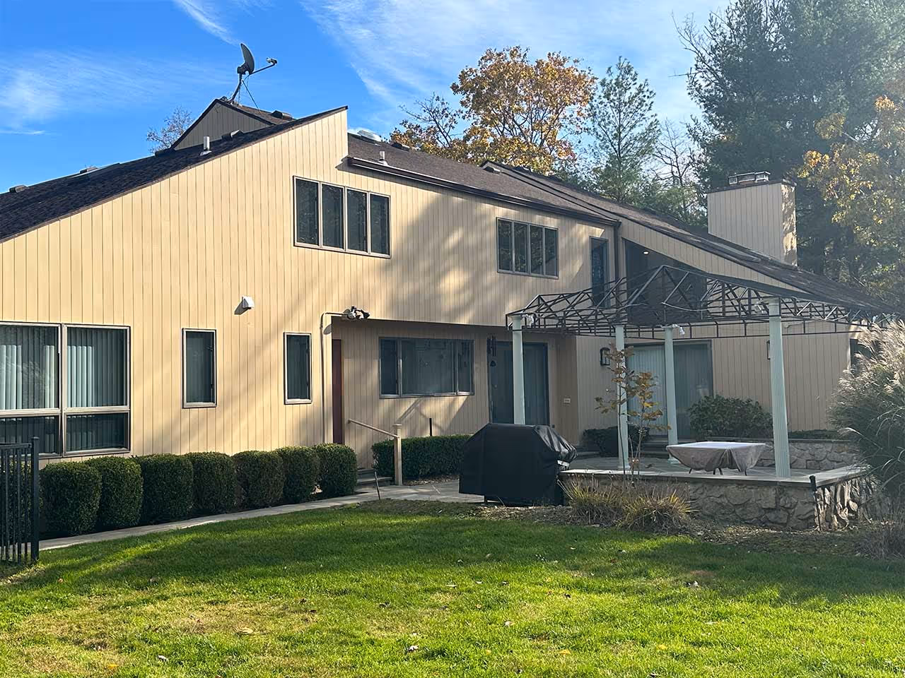 Modern beige house with vertical siding, multiple windows, patio area with metal pergola frame, and green lawn.