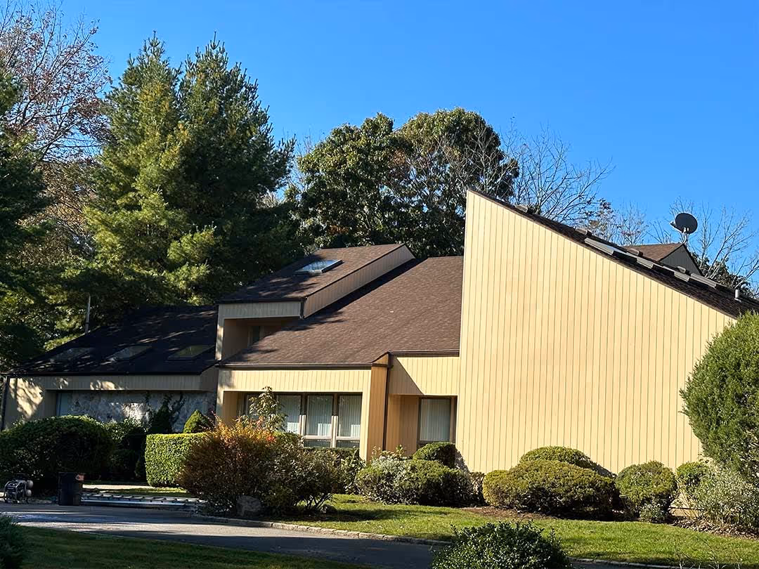 Modern house with beige vertical siding, brown angled roof, surrounded by green bushes and trees under blue sky.