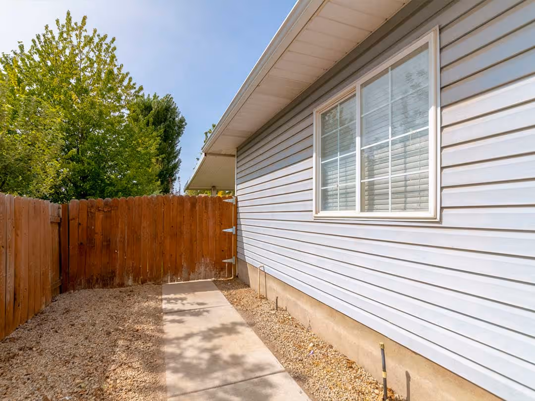 Side view of a house with light gray vinyl siding, a window, gravel ground, concrete path, and wooden fence.