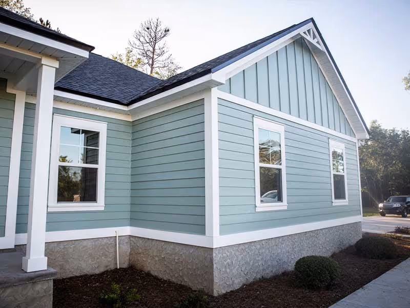 Light blue house exterior with white trim, three windows, dark roof, and stone foundation.