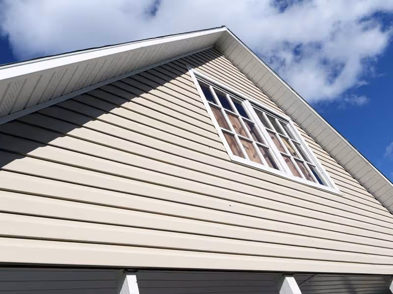 Close-up of beige vinyl siding on a house under a partly cloudy blue sky.