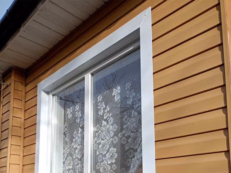 White-framed window with floral lace curtain on a house with beige vinyl siding.