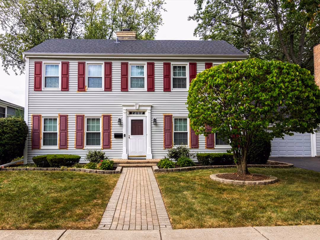 Two-story house with gray vinyl siding, maroon shutters, a central white door, and a green tree in front yard.
