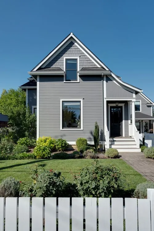 Gray two-story house with white trim, a black door, front porch, manicured lawn, shrubs, and a white picket fence.
